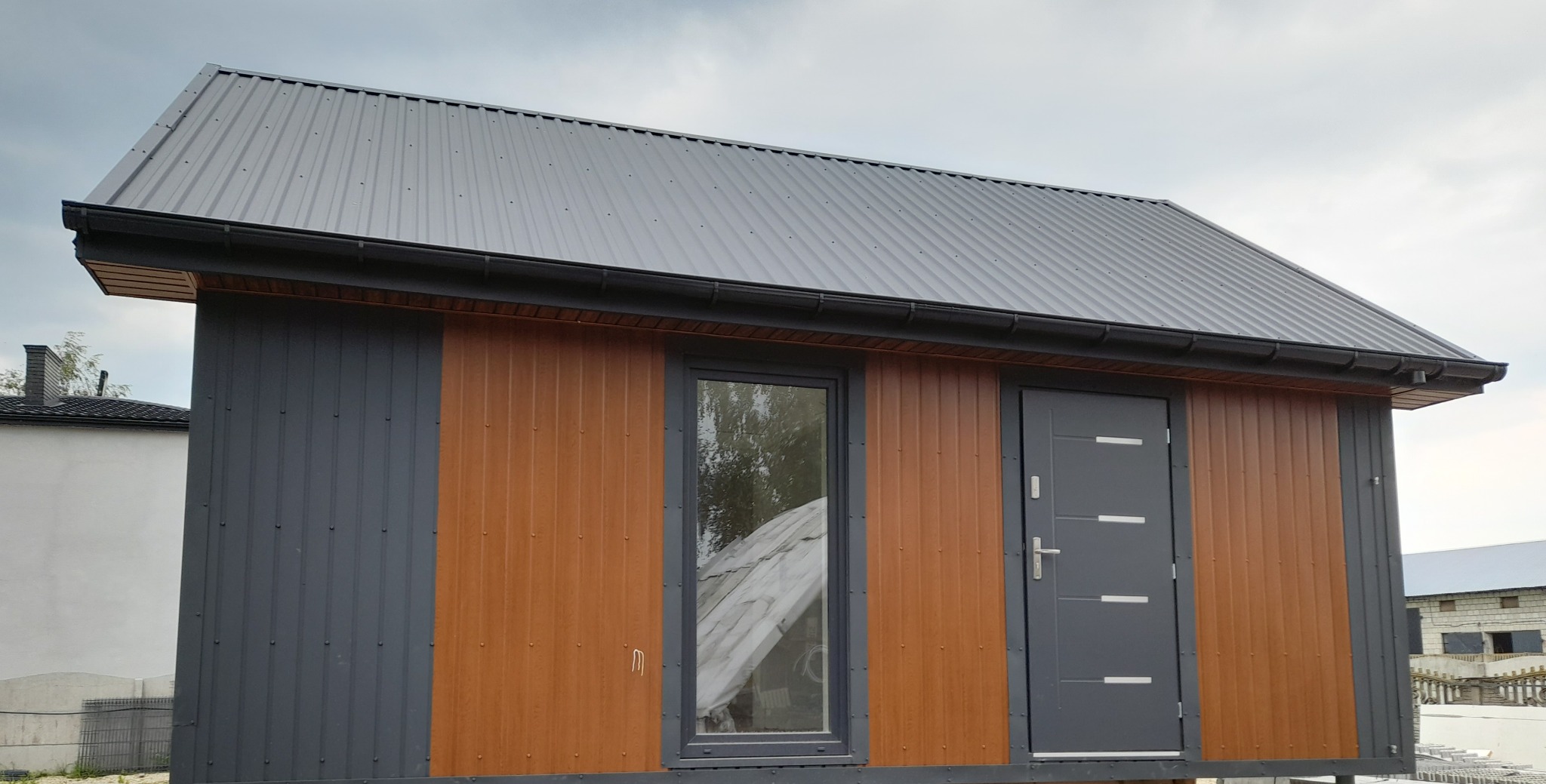 Gotowy, mały domek z szarym dachem z blachy trapezowej, szarymi panelami oraz brązowymi panelami, z szarymi drzwiami i oknem, widok z zewnątrz.