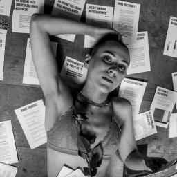 Henryk Szymański Fotografia - Czarno-białe, artystyczne zdjęcie kobiety leżącej na podłodze, otoczonej kartkami z tekstami. Na piersi róża, na szyi łańcuch. Wyraziste oświetlenie i nietypowa kompozycja.