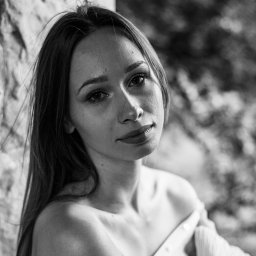 Henryk Szymański Fotografia - Portret młodej kobiety w czerni i bieli, oparta o kamienną ścianę, z delikatnym uśmiechem i długimi włosami, uchwycony w naturalnym świetle.