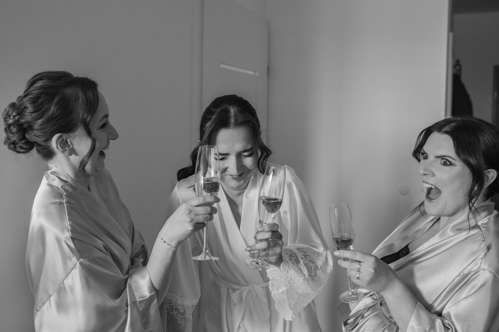 Trzy uśmiechnięte kobiety w szlafrokach wznoszą toast szampanem. Czarno-białe zdjęcie z przygotowań do ślubu, z detalami koronkowych rękawów i biżuterii.