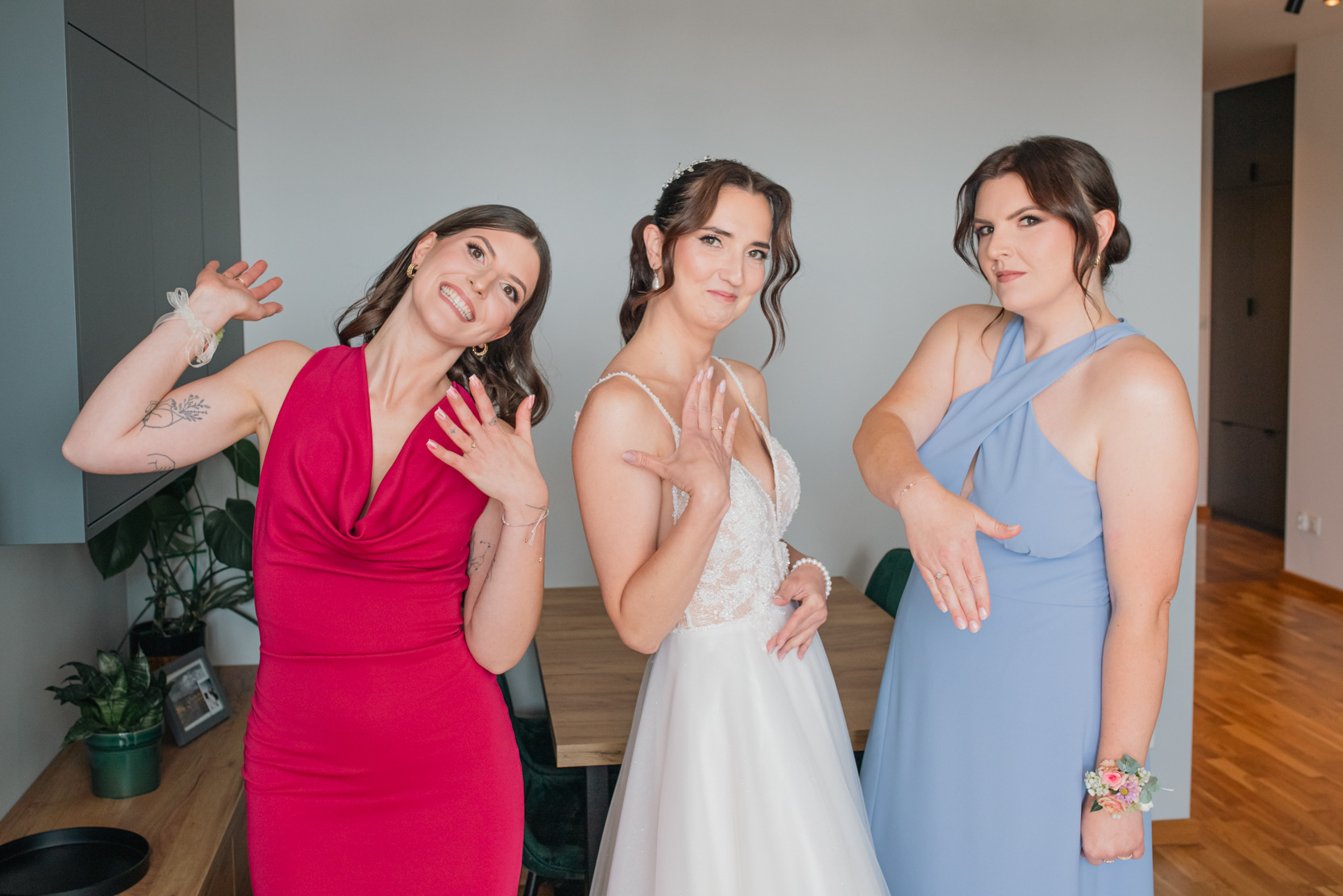 Panna młoda w koronkowej sukni i druhny w kolorowych sukienkach prezentują pierścionki zaręczynowe na tle jasnego wnętrza.