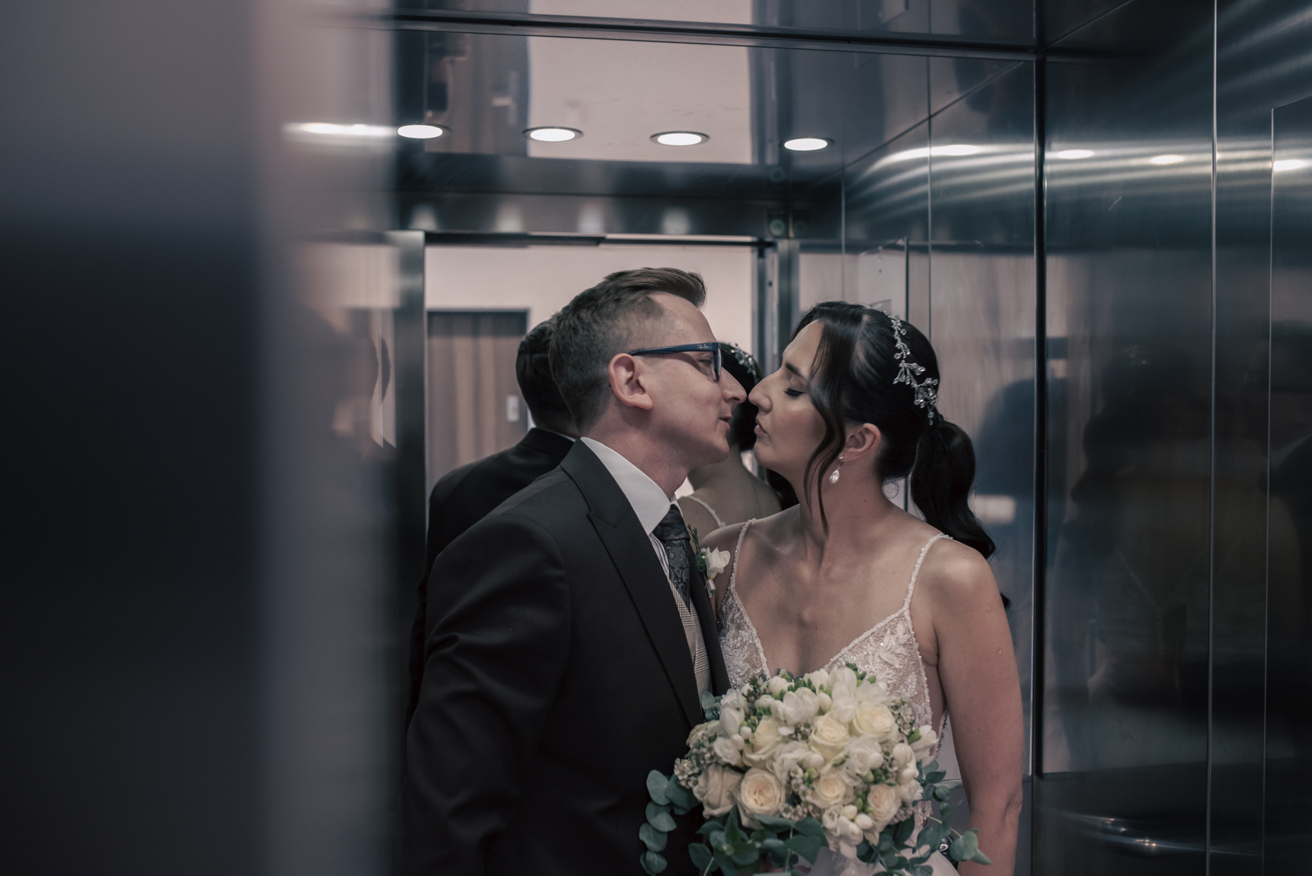 Para młoda w windzie, zbliżająca się do pocałunku. Ona w sukni z bukietem, on w garniturze i okularach. Odbicie w lustrzanych ścianach windy.