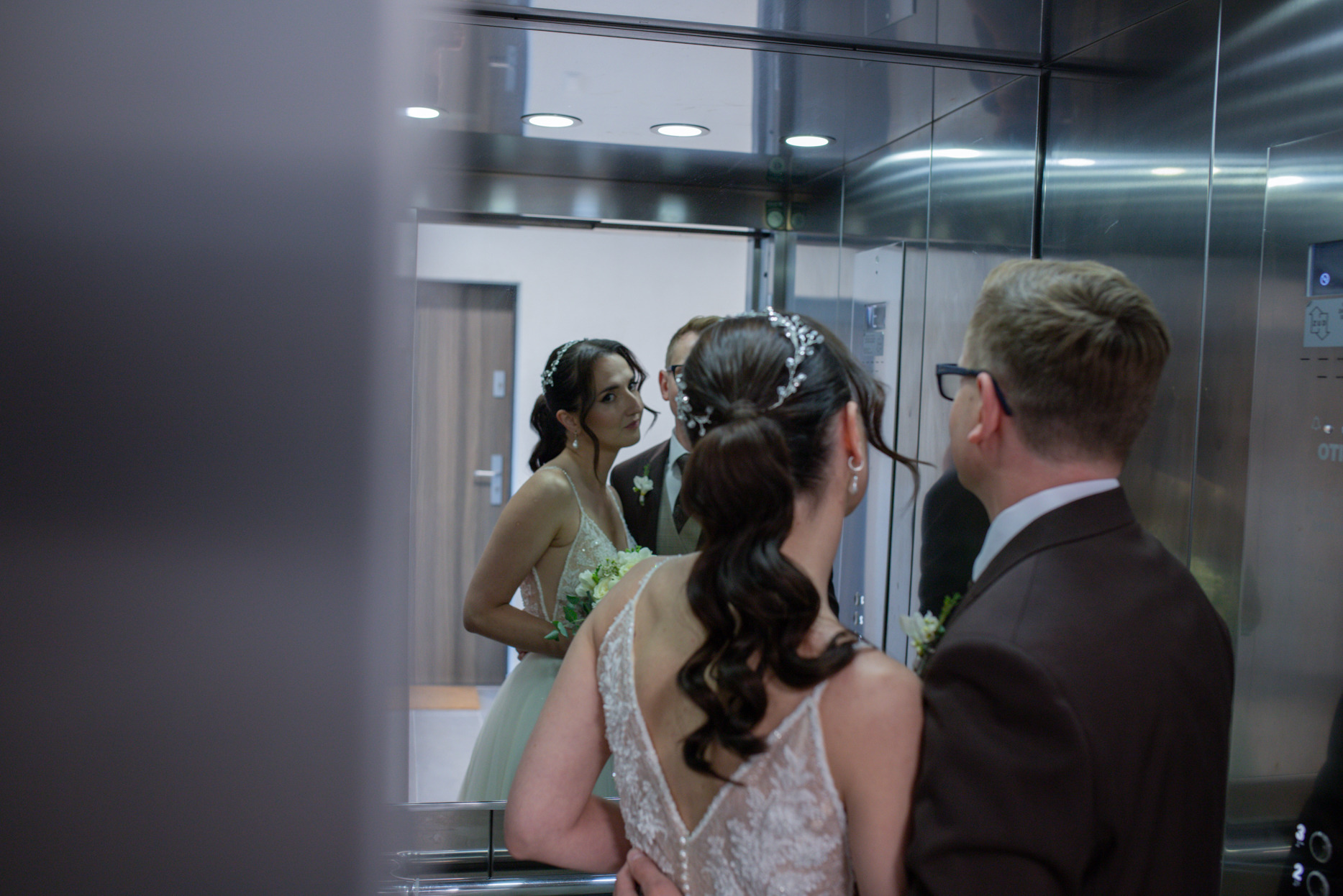 Para młoda w windzie, panna młoda spogląda w lustro. Widoczny fragment sukni ślubnej z koronkowym wykończeniem i upięcie włosów. Pan młody w brązowym garniturze.