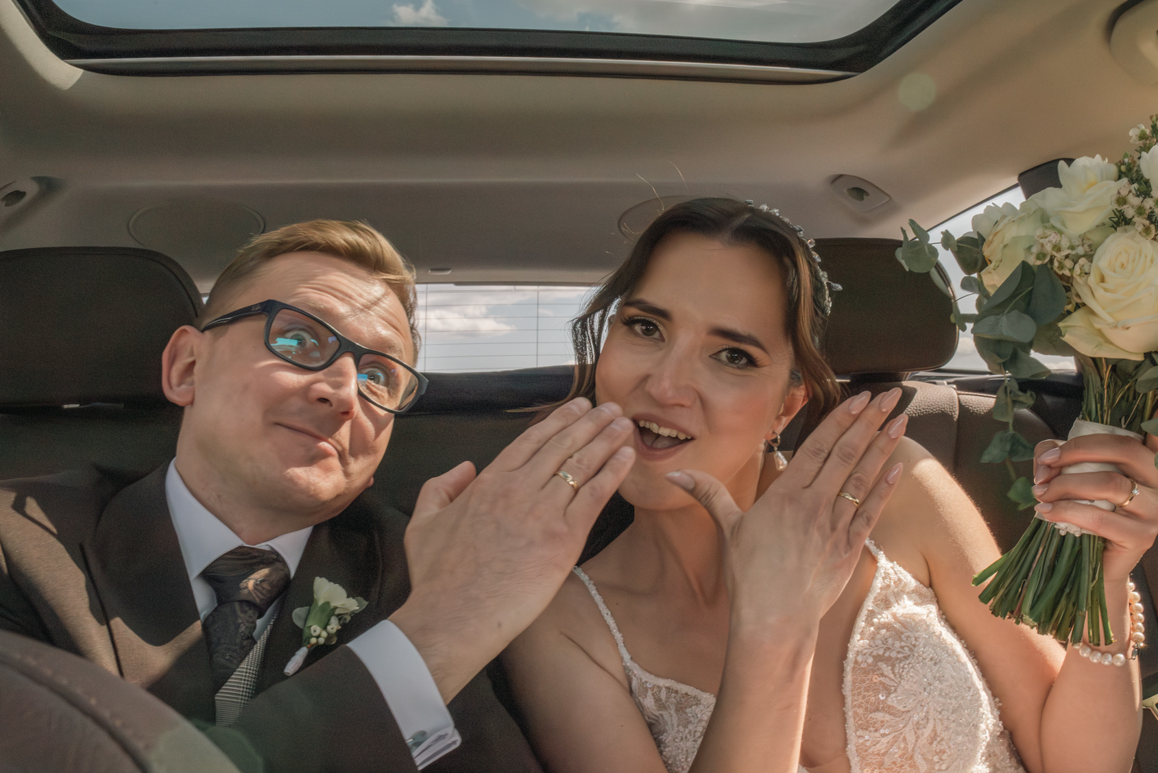 Uśmiechnięta para młoda w samochodzie z otwartym dachem, prezentująca obrączki. Panna młoda trzyma bukiet białych róż. Radosne ujęcie z dnia ślubu.