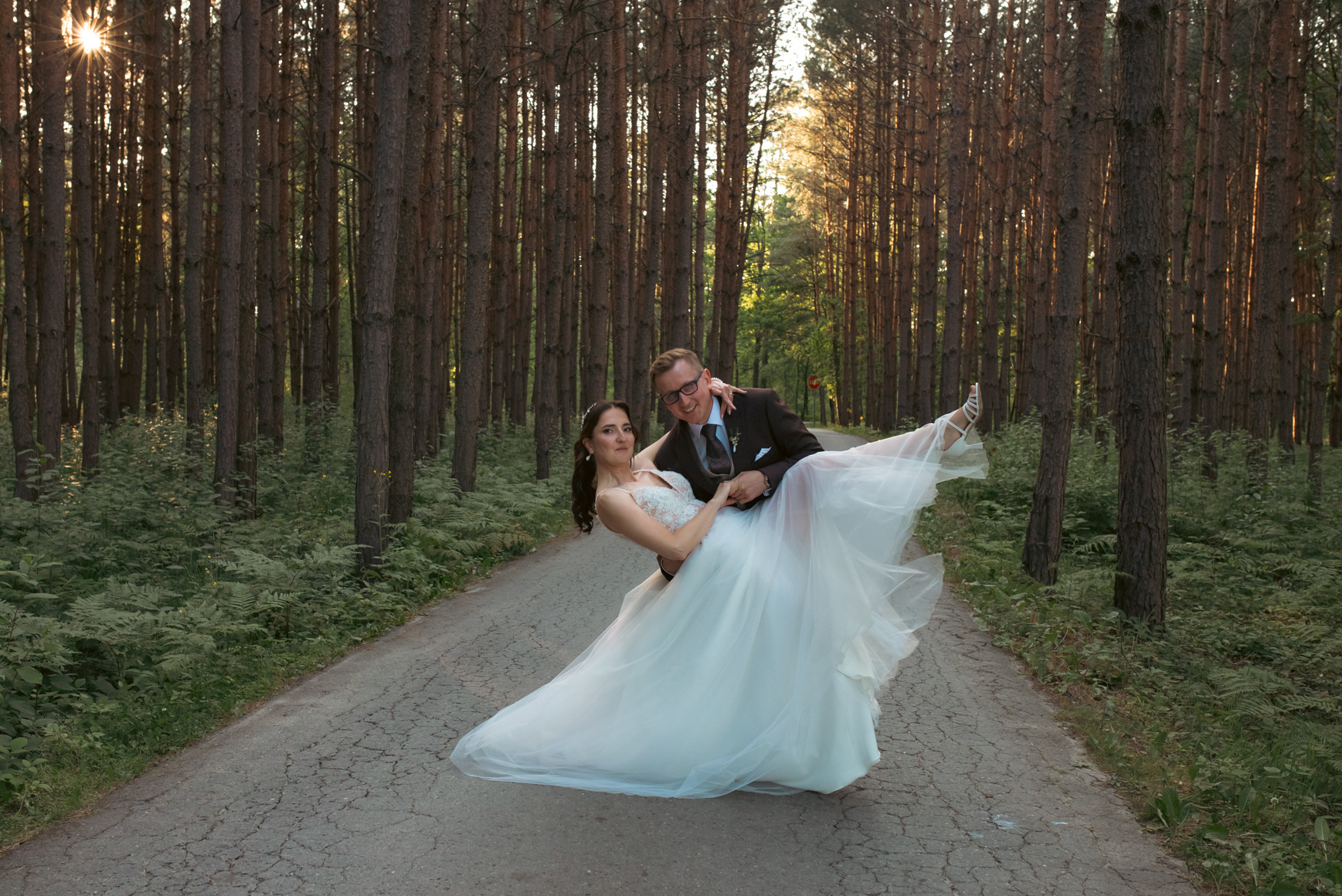 Uśmiechnięty pan młody trzyma pannę młodą w białej sukni na tle leśnej drogi, promienie słońca przebijają przez drzewa. Romantyczna sesja ślubna w plenerze.