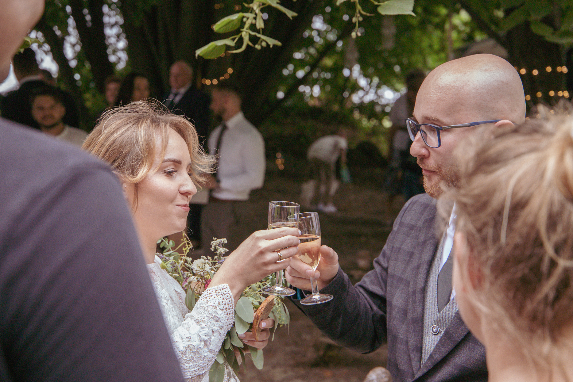 Para młoda wznosi toast szampanem na tle zieleni, widać obrączkę na palcu panny młodej, mężczyzna w garniturze w kratę i okularach, uśmiechnięta kobieta w białej sukni.