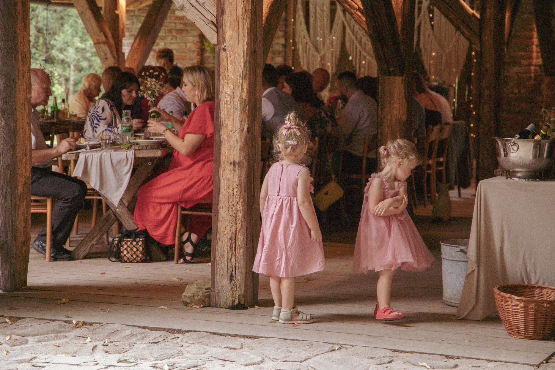 Dwie małe dziewczynki w różowych sukienkach na tle rustykalnego przyjęcia weselnego z drewnianymi elementami i gośćmi przy stołach.