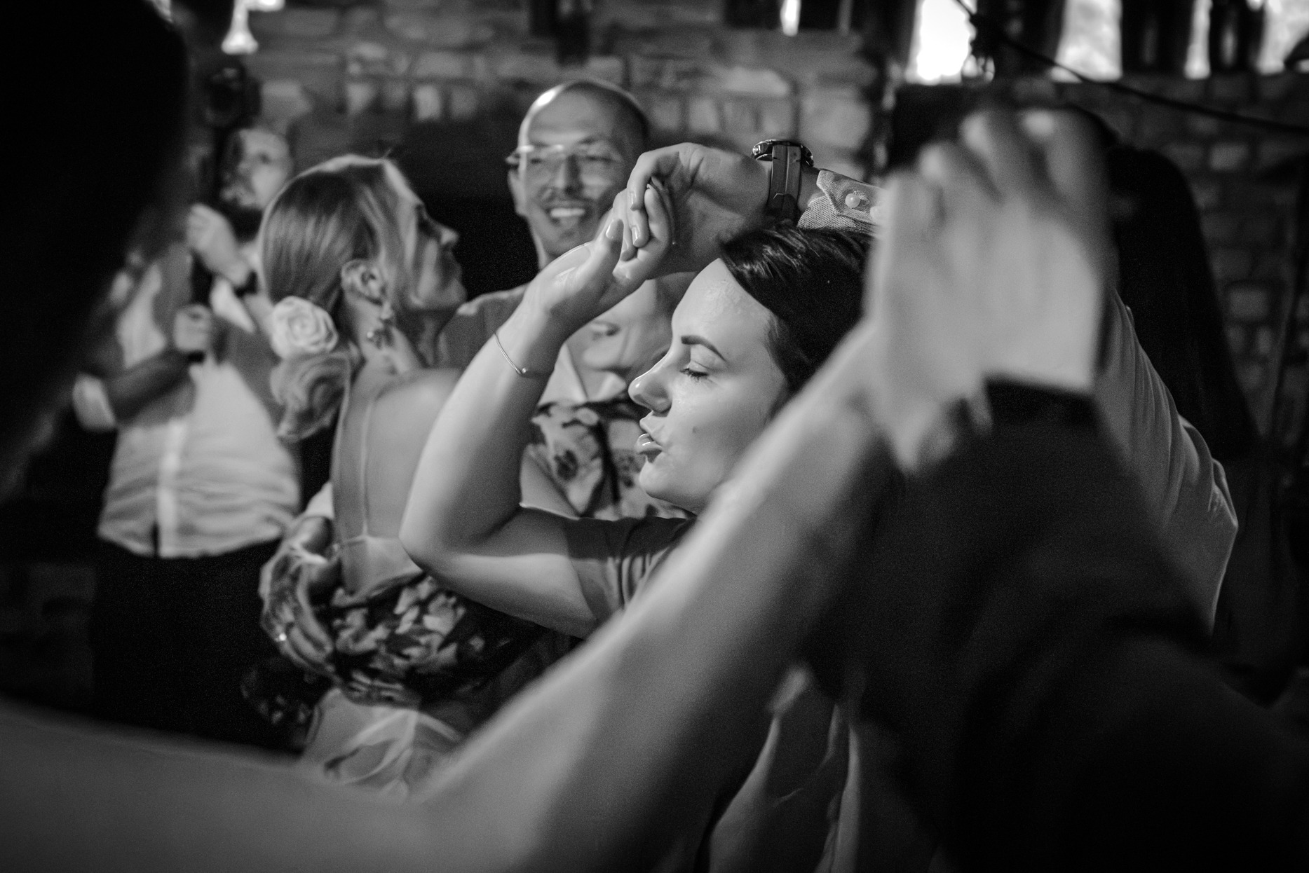 Czarno-białe zdjęcie z wesela: panna młoda z zamkniętymi oczami, trzymająca ręce w górze, w tle uśmiechnięty pan młody i goście. Dynamiczne ujęcie z parkietu.