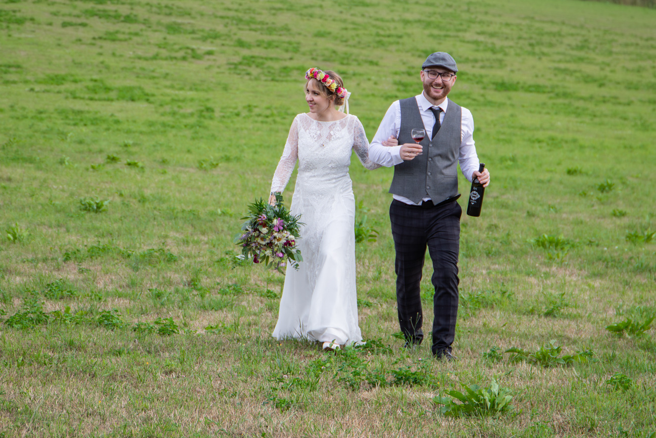Uśmiechnięta para młoda spaceruje po zielonej łące. Panna młoda w koronkowej sukni trzyma bukiet, pan młody w kamizelce i czapce niesie wino i kieliszek.