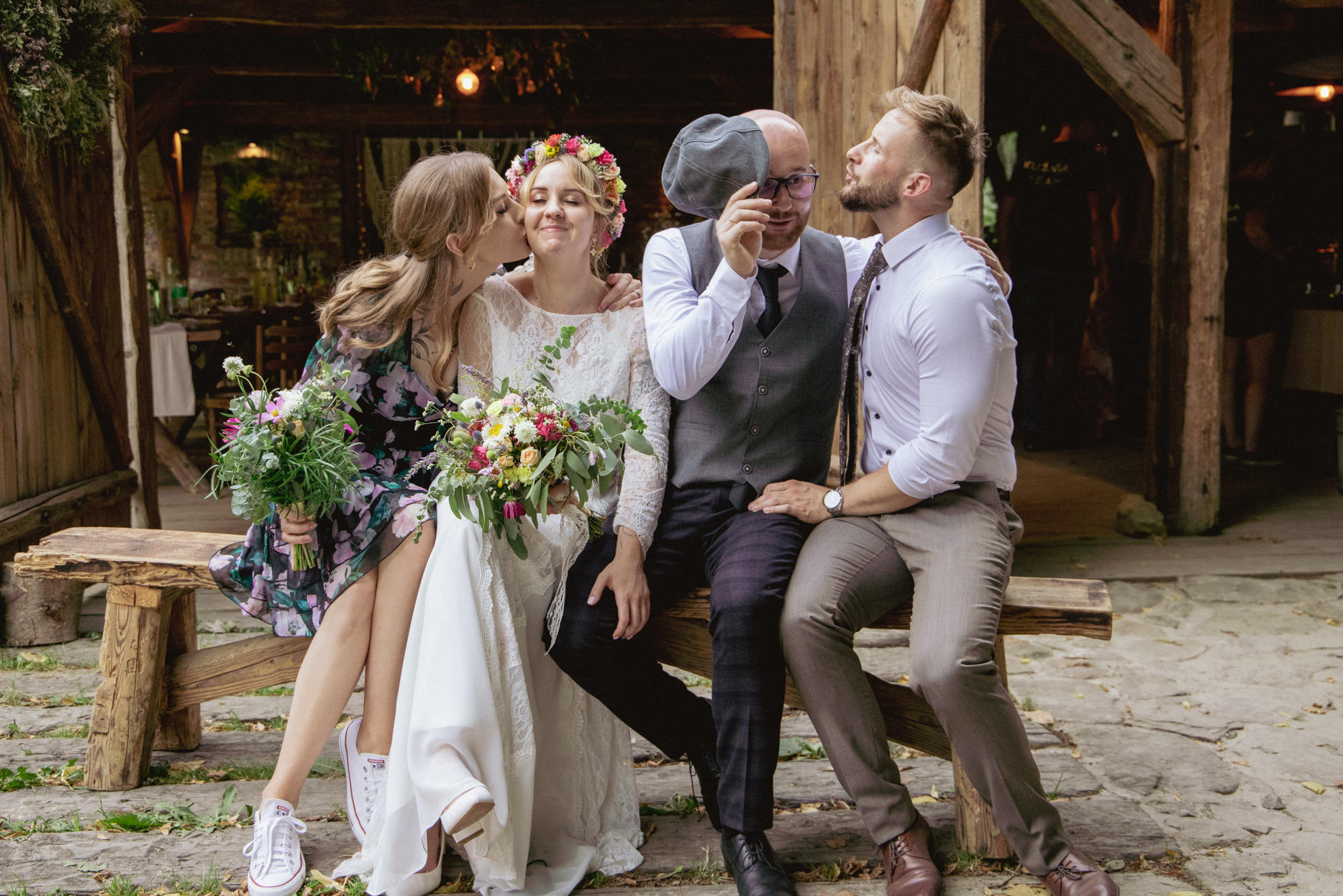 Uśmiechnięta panna młoda w wianku, całowana przez przyjaciół na rustykalnym tle. Nietypowe ujęcie z sesji ślubnej, z humorem i naturalnością.