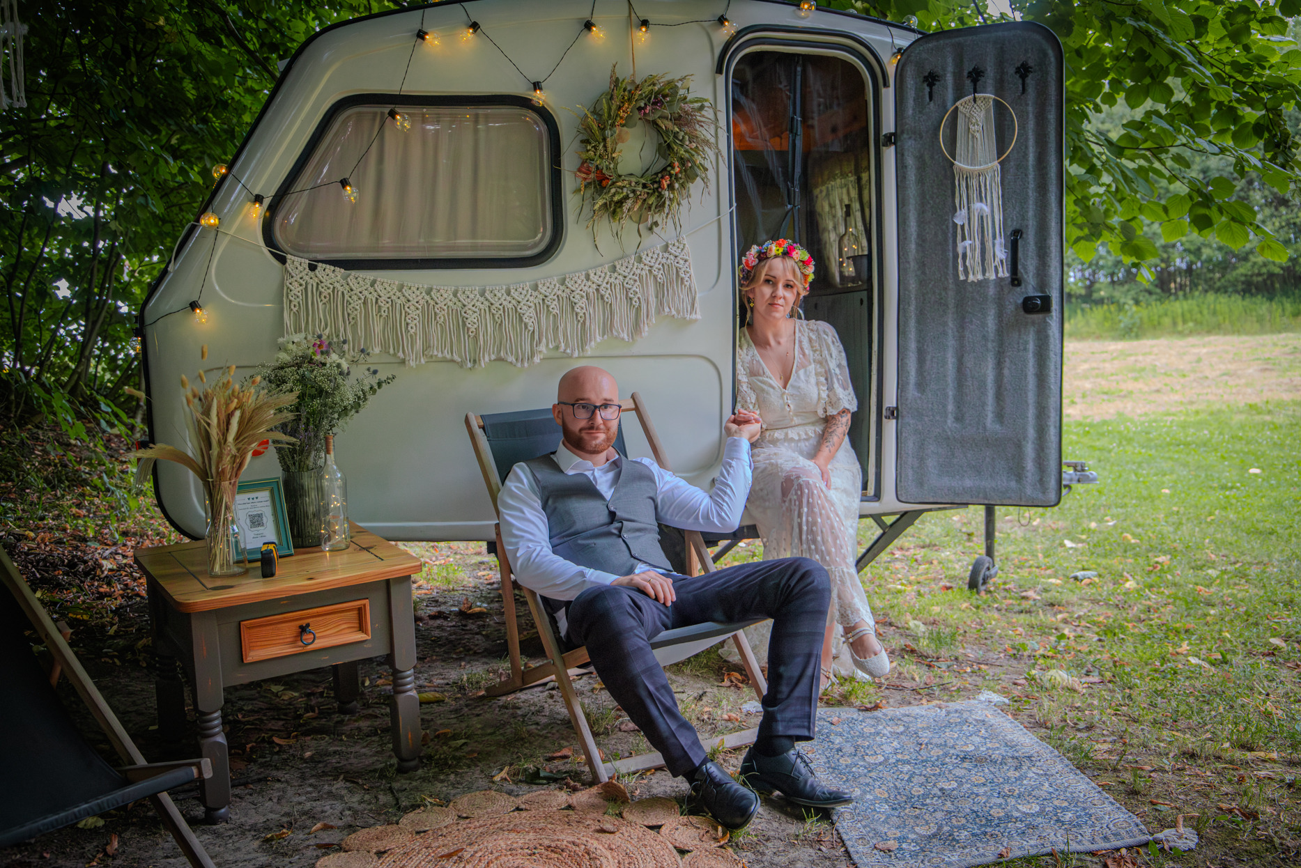 Para młoda pozuje przy dekorowanym kamperze w stylu boho. Panna młoda w wianku i koronkowej sukni, pan młody w garniturze. Romantyczna sesja plenerowa.