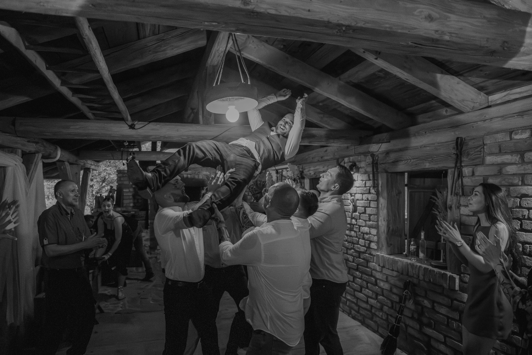 Czarno-białe zdjęcie z wesela: grupa mężczyzn podrzuca w górę uśmiechniętego pana młodego w rustykalnym wnętrzu z drewnianym stropem i ceglanymi ścianami. Goście klaszczą.