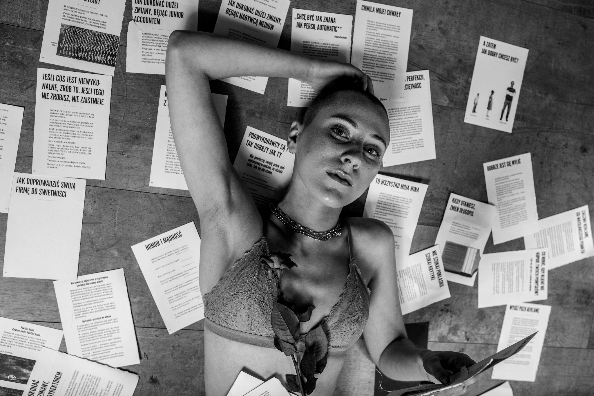 Czarno-białe, artystyczne zdjęcie kobiety leżącej na podłodze, otoczonej kartkami z tekstami. Na piersi róża, na szyi łańcuch. Wyraziste oświetlenie i nietypowa kompozycja.