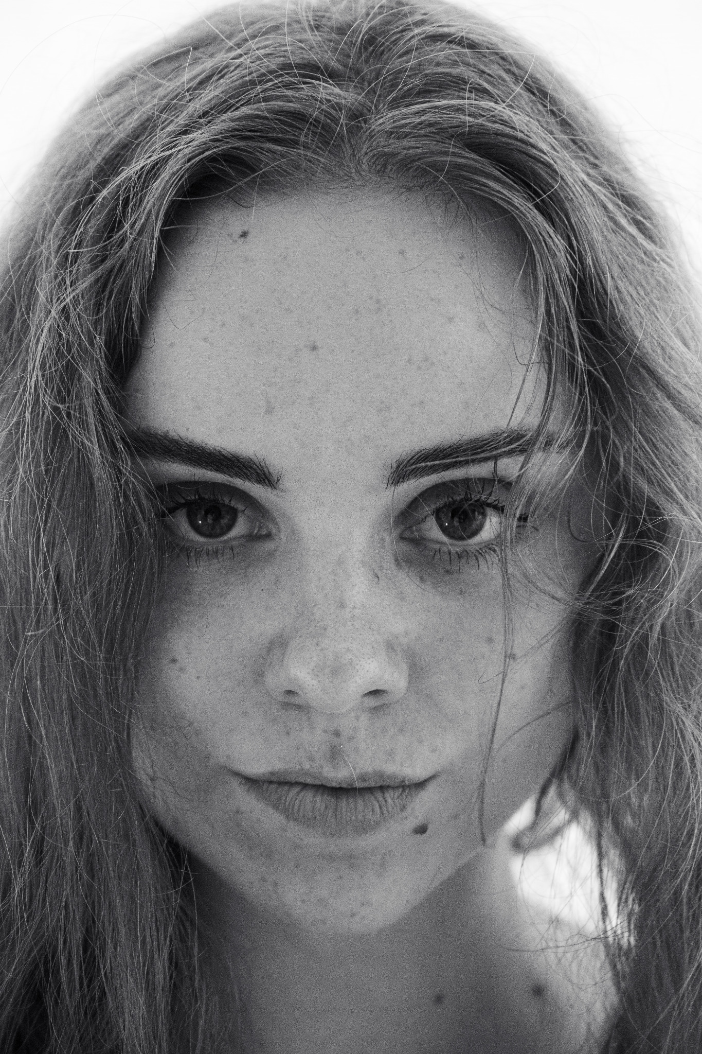 Czarno-biały portret kobiety z bliska, z widocznymi piegami i rozpuszczonymi włosami, skierowana prosto w obiektyw, z intensywnym spojrzeniem.