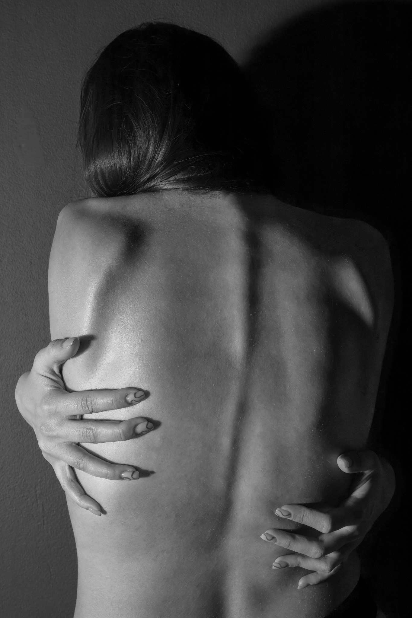 Monochromatyczne ujęcie pleców kobiety obejmującej się rękoma, z delikatnym wzorem na paznokciach, na tle ciemnej ściany. Subtelne światłocienie podkreślają anatomię.