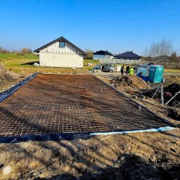 DOM PROJEKT STABILNY FUNDAMENT sp. z o.o - Przygotowanie zbrojenia pod płytę fundamentową na działce budowlanej z widocznym budynkiem w stanie surowym otwartym, przenośną toaletą i grupą osób w tle.