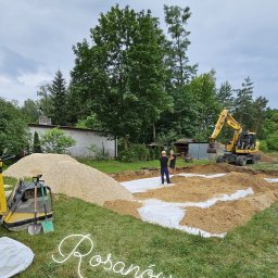 DOM PROJEKT STABILNY FUNDAMENT sp. z o.o - Żółta koparka Komatsu podczas prac ziemnych przy budowie fundamentów, widoczne dwie osoby nadzorujące prace, stos piasku i zagęszczarka obok wykopu.