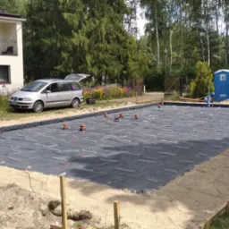 Przygotowany teren pod budowę z rozłożoną folią izolacyjną, wystającymi rurami wentylacyjnymi i widocznym szarym samochodem osobowym w tle.