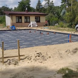 DOM PROJEKT STABILNY FUNDAMENT sp. z o.o - Przygotowanie podłoża pod fundamenty: czarna folia izolacyjna z białymi elementami dystansowymi, wystające rury kanalizacyjne, piasek, w tle dom jednorodzinny i zaparkowany samochód.