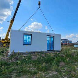 DOM PROJEKT STABILNY FUNDAMENT sp. z o.o - Żółty dźwig podnosi gotowy moduł domu z dwoma oknami zabezpieczonymi folią, widoczna trawa i inne budynki w tle.