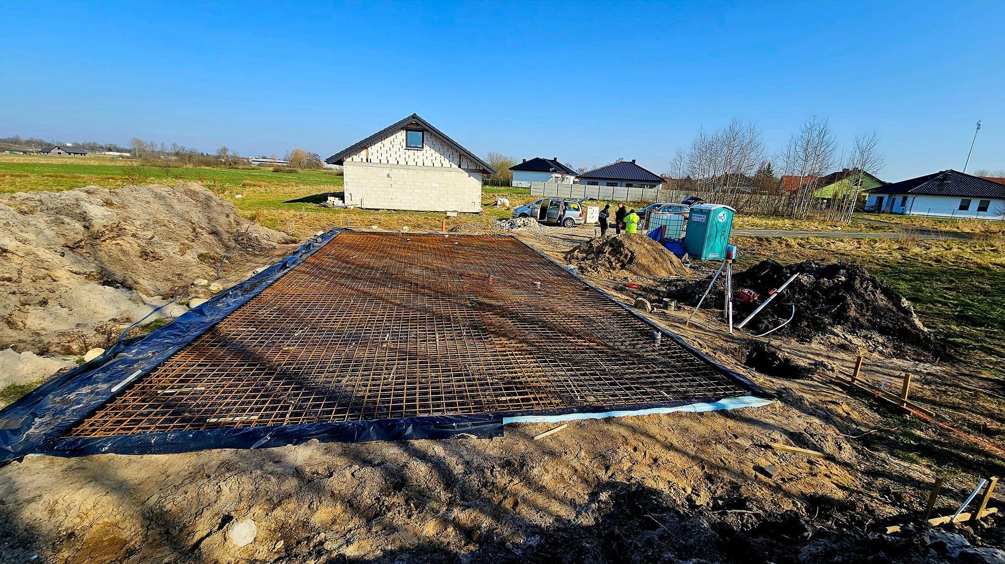 Przygotowanie zbrojenia pod płytę fundamentową na działce budowlanej z widocznym budynkiem w stanie surowym otwartym, przenośną toaletą i grupą osób w tle.