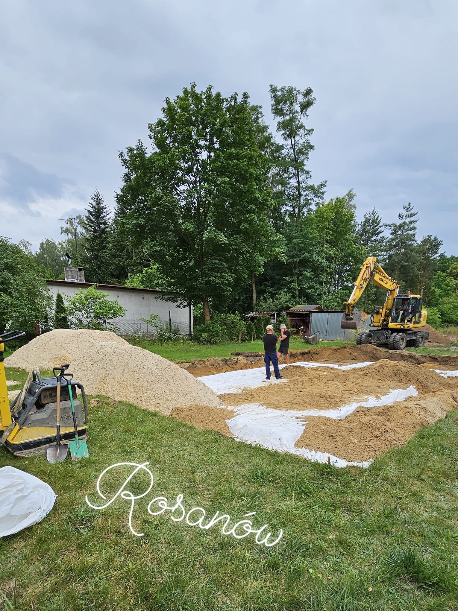 Żółta koparka Komatsu podczas prac ziemnych przy budowie fundamentów, widoczne dwie osoby nadzorujące prace, stos piasku i zagęszczarka obok wykopu.