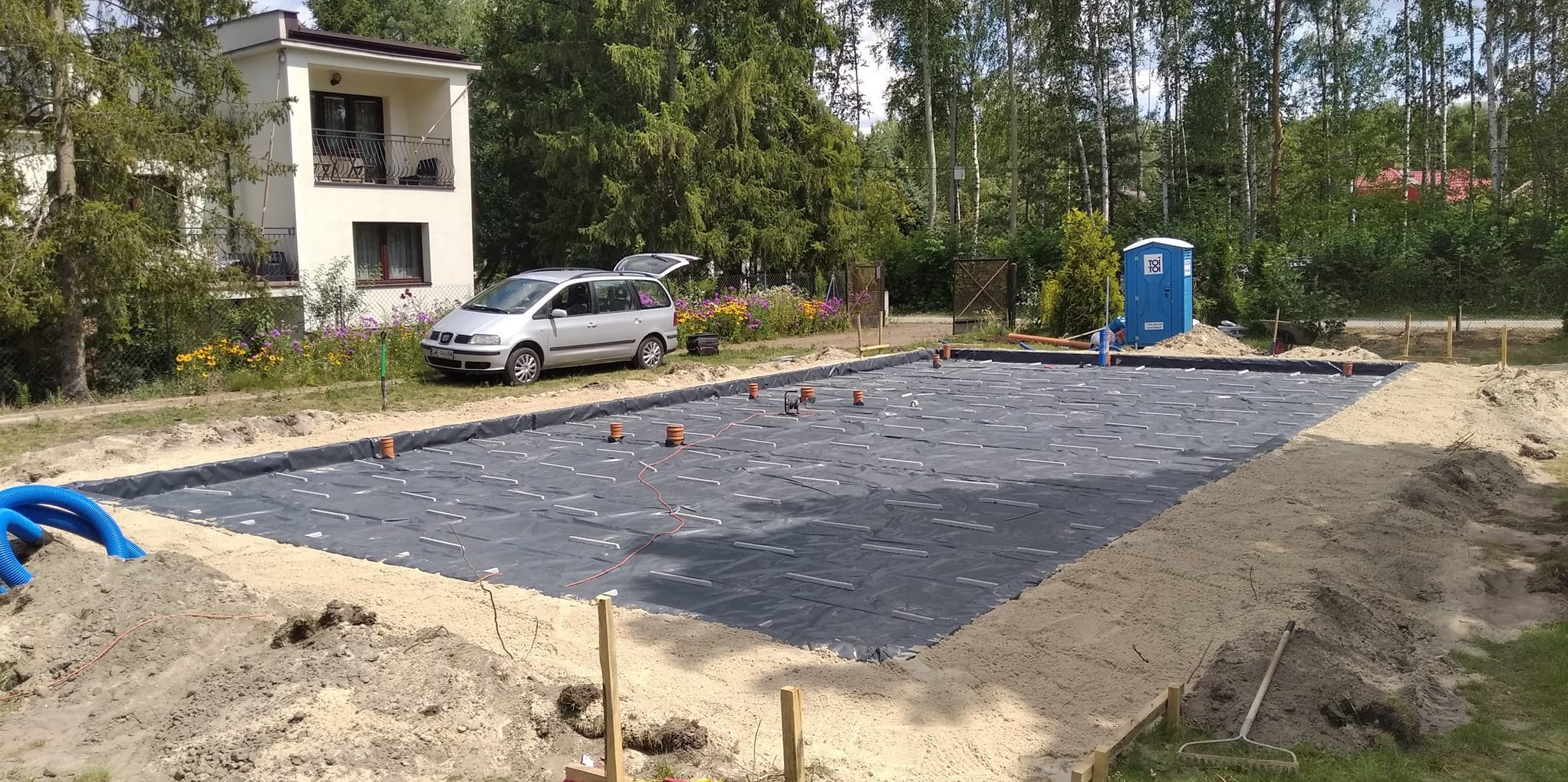 Przygotowany teren pod budowę z rozłożoną folią izolacyjną, wystającymi rurami wentylacyjnymi i widocznym szarym samochodem osobowym w tle.