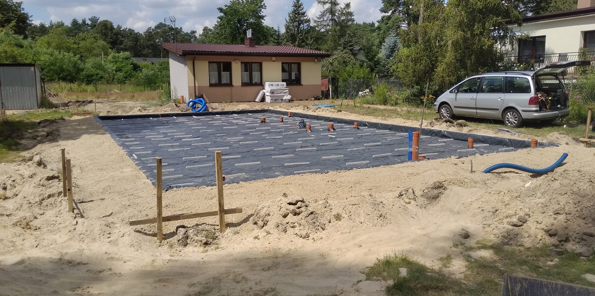 Przygotowanie podłoża pod fundamenty: czarna folia izolacyjna z białymi elementami dystansowymi, wystające rury kanalizacyjne, piasek, w tle dom jednorodzinny i zaparkowany samochód.