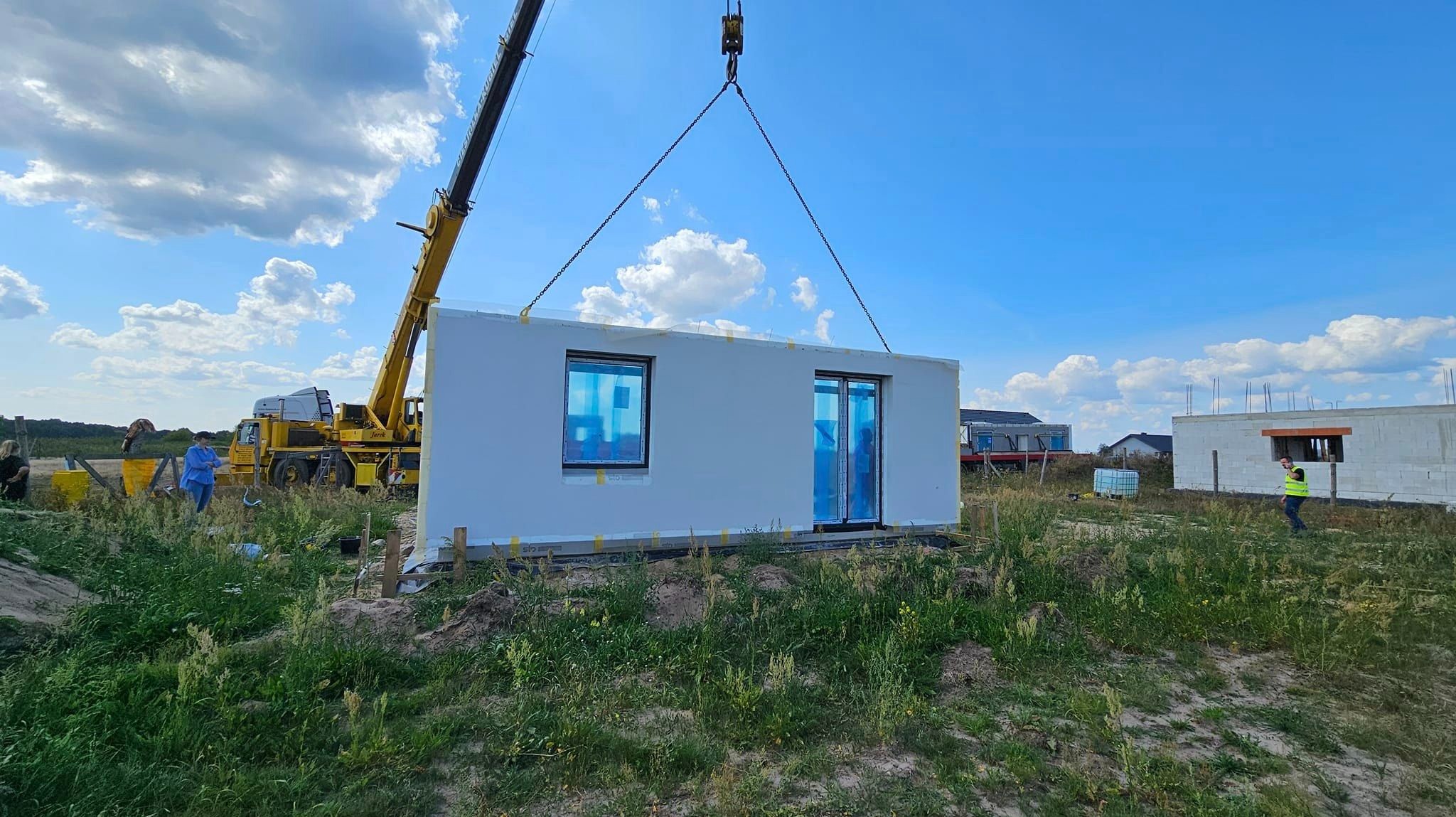 Żółty dźwig podnosi gotowy moduł domu z dwoma oknami zabezpieczonymi folią, widoczna trawa i inne budynki w tle.