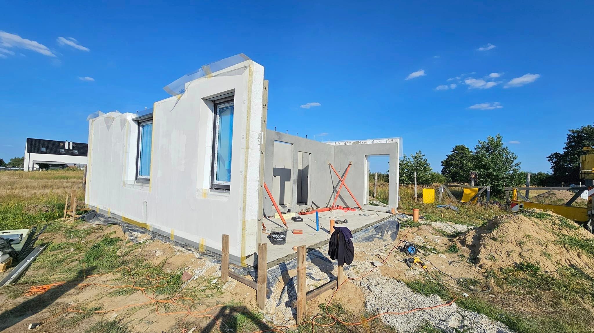 Wznoszenie szkieletowej konstrukcji domu z prefabrykatów, widoczne ściany z oknami, stropy, elementy mocujące i narzędzia na placu budowy w słoneczny dzień.