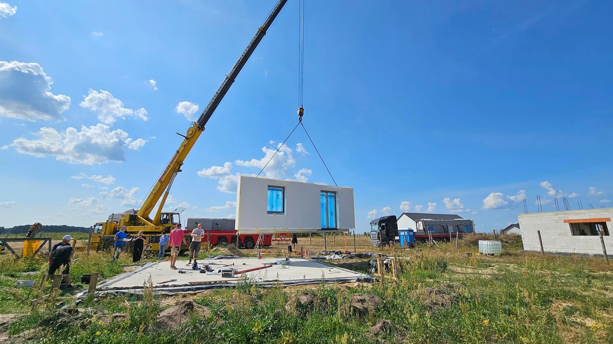 Montaż modułowego domu z użyciem dźwigu na przygotowanym fundamencie, widoczni pracownicy budowlani i ciężarówka na tle błękitnego nieba.