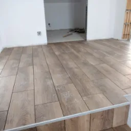 Ułożona podłoga z płytek imitujących drewno, widoczny próg i fragment ściany z puszkami elektrycznymi, w tle pomieszczenie w stanie surowym.