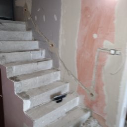 Jakub Łukomski - Schody betonowe w trakcie remontu, widoczne ślady gładzi na ścianach, kabel wystający ze ściany, szpachelka na schodach, wiadro na podłodze.