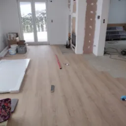 Układanie paneli podłogowych w jasnym pomieszczeniu z dużym oknem balkonowym, widoczne narzędzia, poziomica, paczka paneli i fragment ściany przygotowanej pod gładzie.