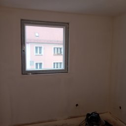 Jakub Łukomski - Pokój w trakcie remontu, ściany przygotowane pod malowanie, widoczny odkurzacz przemysłowy i okno z widokiem na różowy budynek.