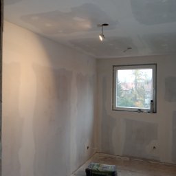 Jakub Łukomski - Pokój w trakcie remontu, ściany pokryte częściowo gładzią, widoczne worki z betonem na podłodze i surowa oprawa oświetleniowa.