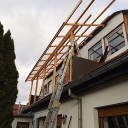 Jakub Łukomski - Montaż drewnianej konstrukcji zadaszenia na balkonie domu jednorodzinnego, widoczna drabina oparta o dach i fragment osoby w dolnym rogu kadru.