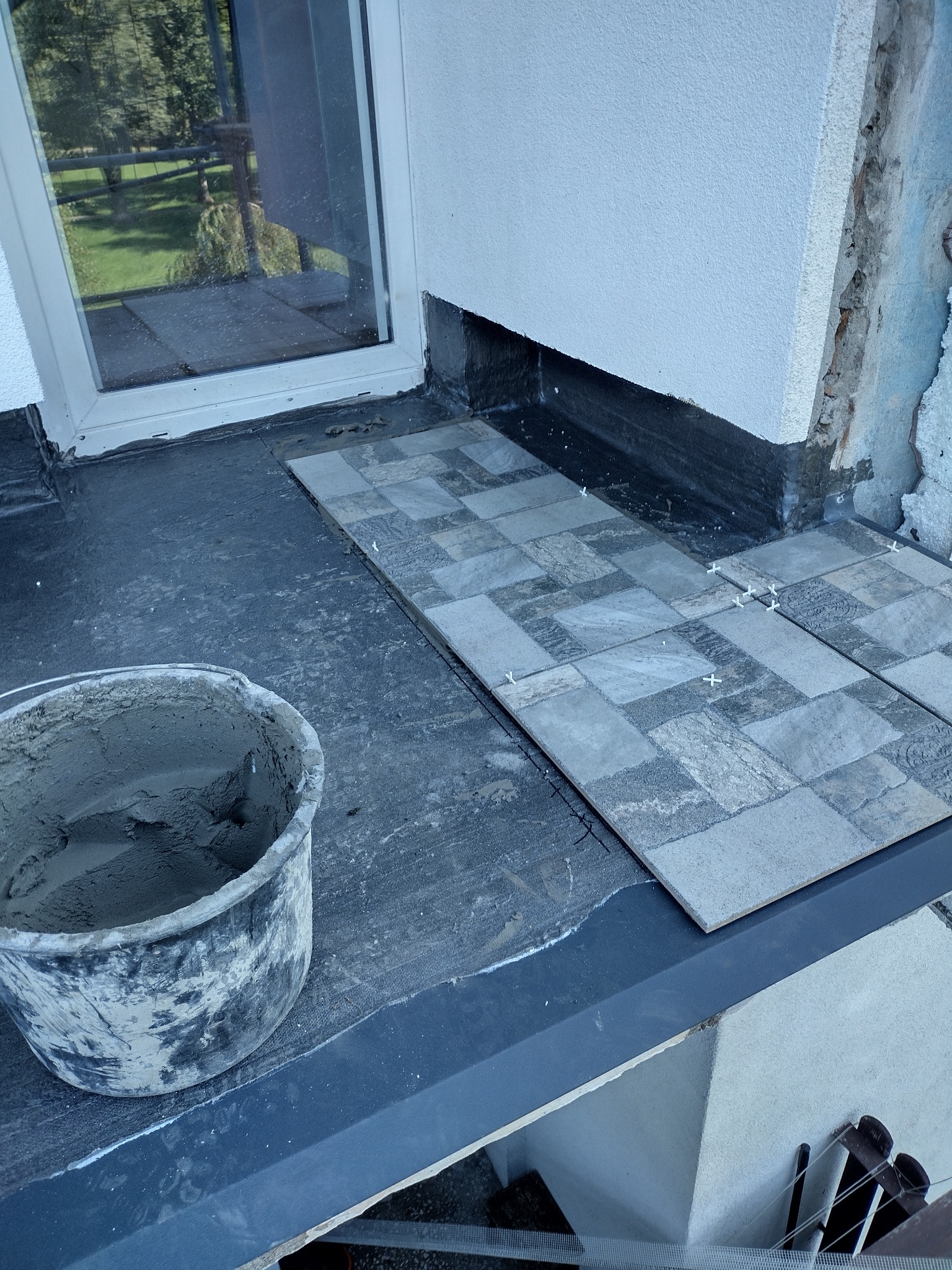 Układanie szaro-beżowych płytek na tarasie z widocznym wiadrem z zaprawą i krzyżykami dystansowymi, fragment ściany z uszkodzonym tynkiem.