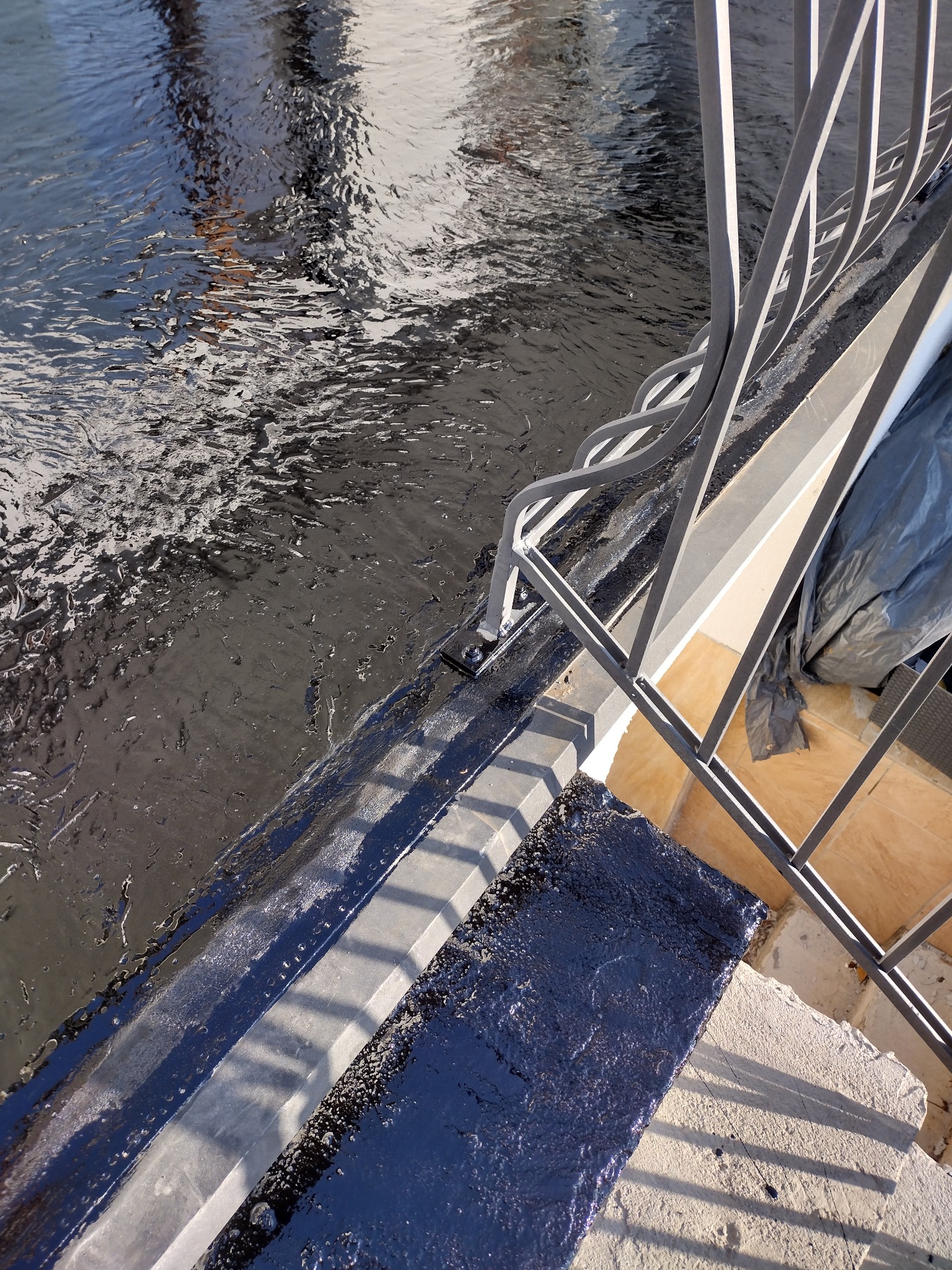 Hydroizolacja balkonu z widoczną, świeżo nałożoną, czarną powłoką bitumiczną, metalową balustradą i fragmentem tarasu w tle.