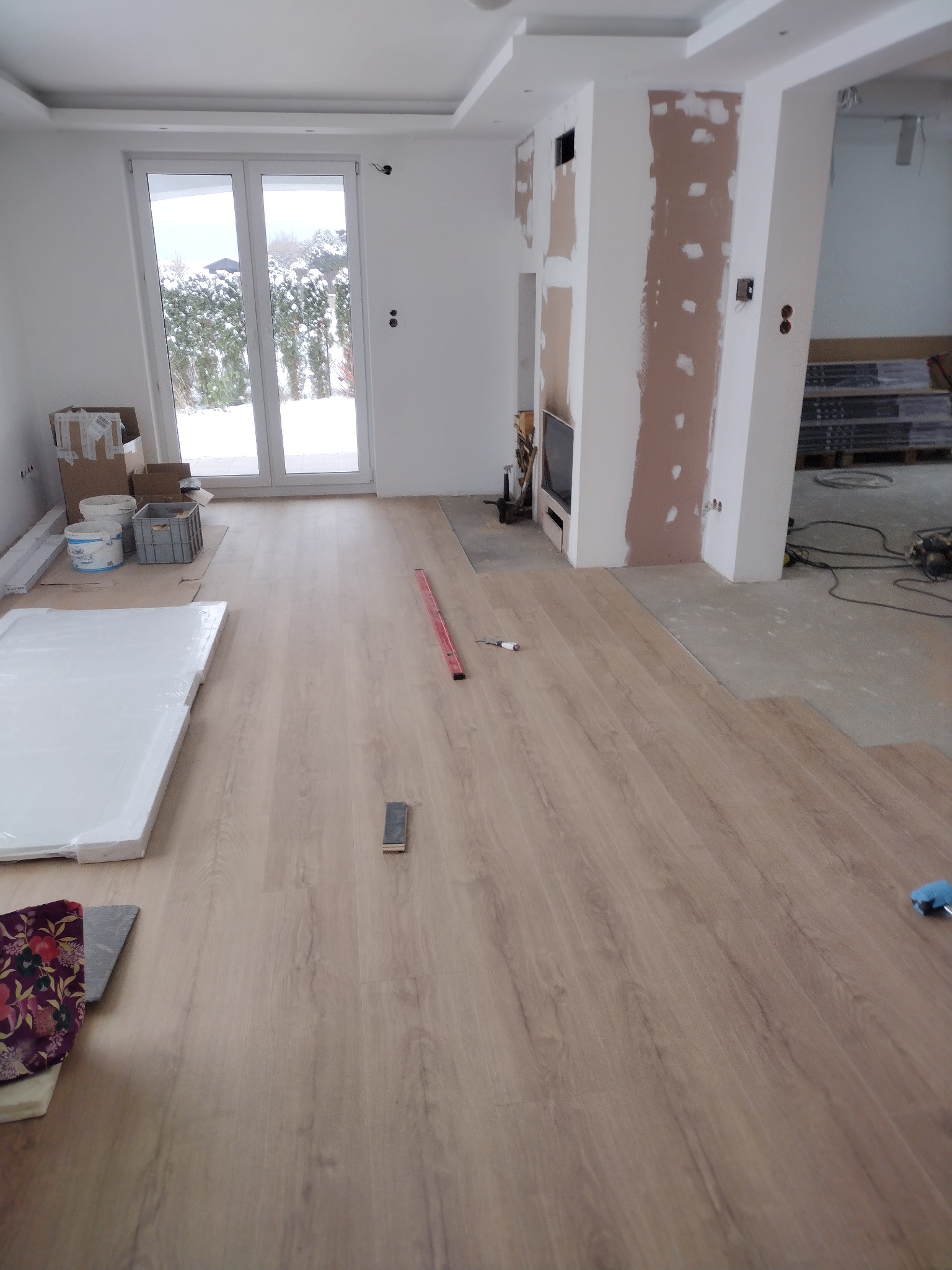 Układanie paneli podłogowych w jasnym pomieszczeniu z dużym oknem balkonowym, widoczne narzędzia, poziomica, paczka paneli i fragment ściany przygotowanej pod gładzie.