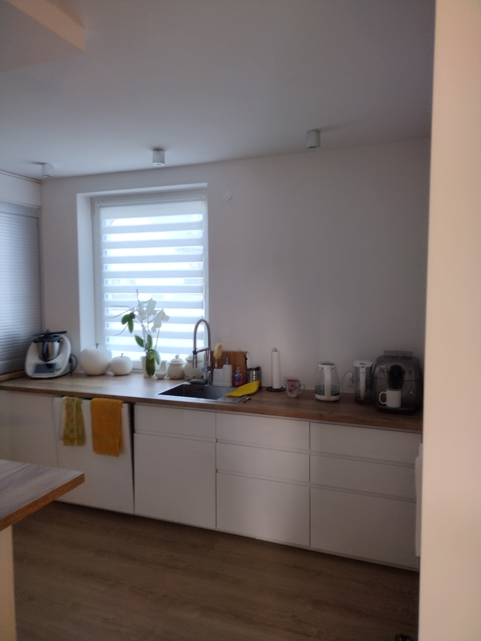 Jasna kuchnia z białymi szafkami i drewnianym blatem, widok na zlewozmywak, okno z żaluzjami i różne sprzęty kuchenne.