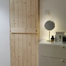 Drewniane drzwi przesuwne w stylu rustykalnym, wykonane z surowych desek, widoczne obok komody z lustrem w oświetlonym pomieszczeniu.