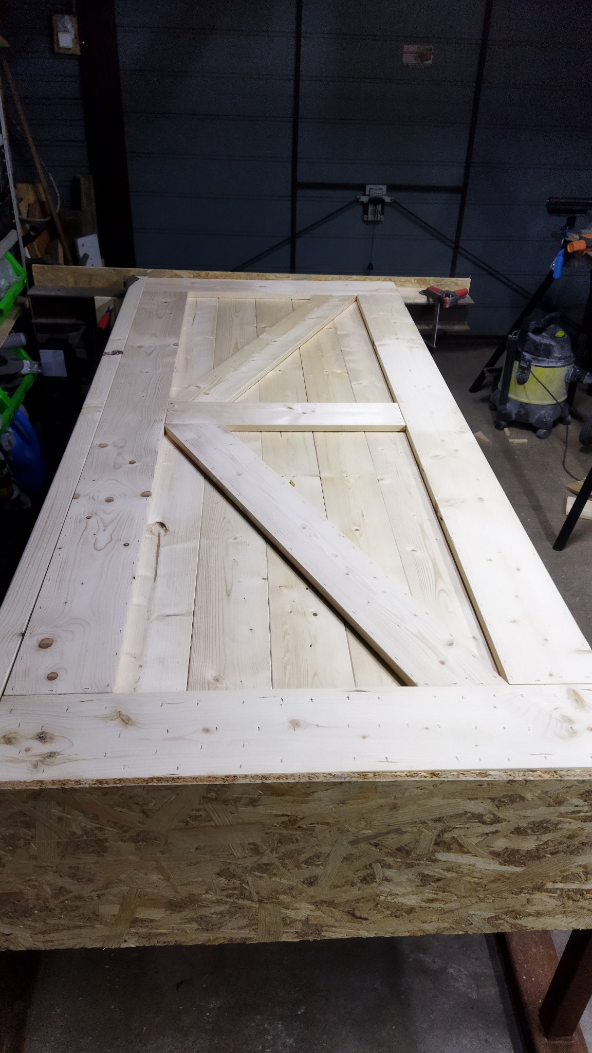 Surowa drewniana brama w stylu rustykalnym z ukośnym wzmocnieniem, leżąca na płycie OSB w warsztacie, widoczne ślady po zszywkach.