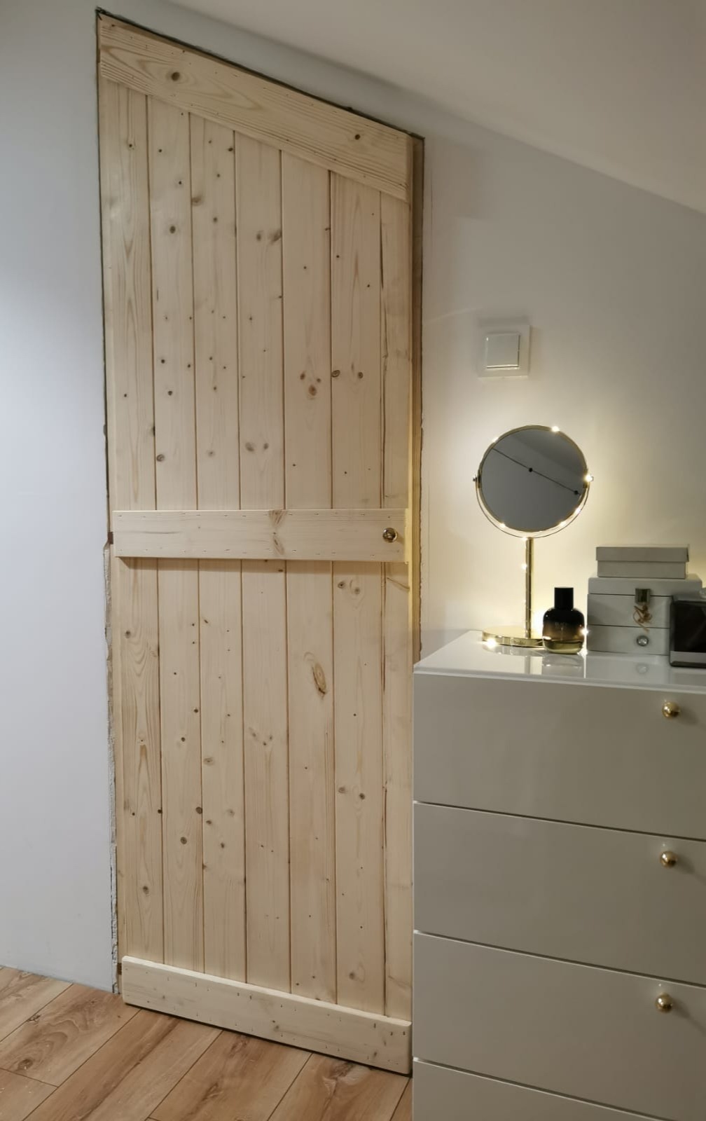 Drewniane drzwi przesuwne w stylu rustykalnym, wykonane z surowych desek, widoczne obok komody z lustrem w oświetlonym pomieszczeniu.