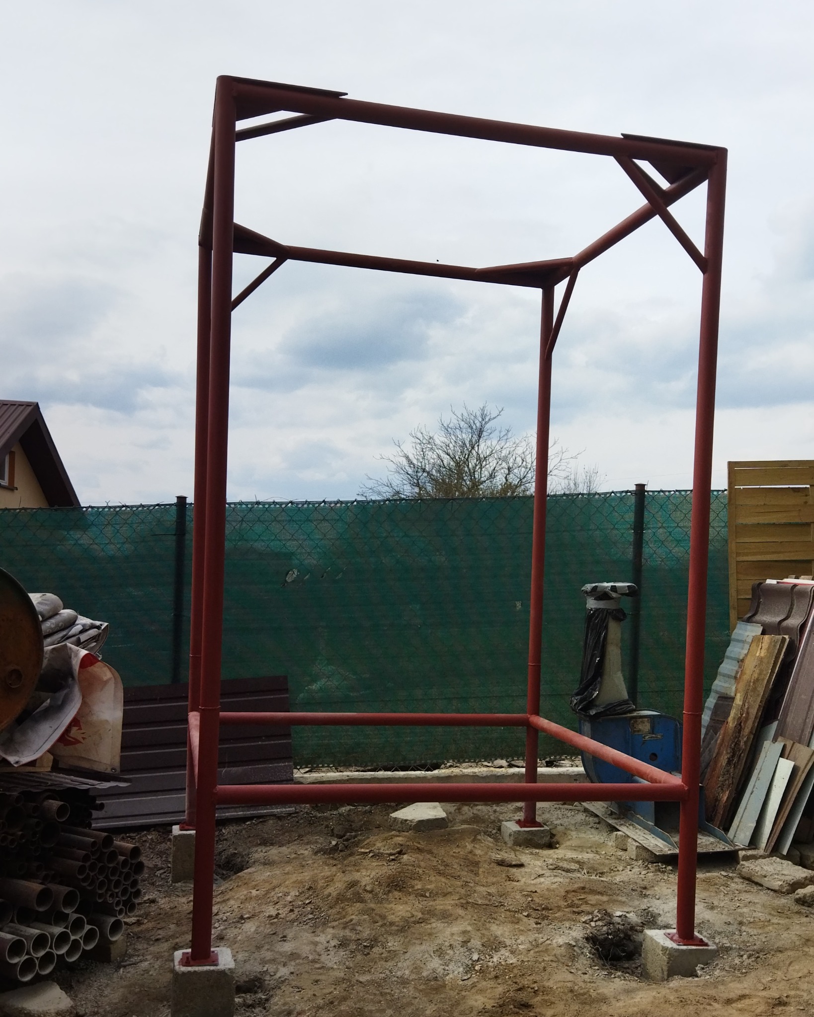 Czerwona, metalowa konstrukcja szkieletowa w trakcie budowy, osadzona na betonowych fundamentach, z widocznym zielonym ogrodzeniem i elementami budowlanymi w tle.