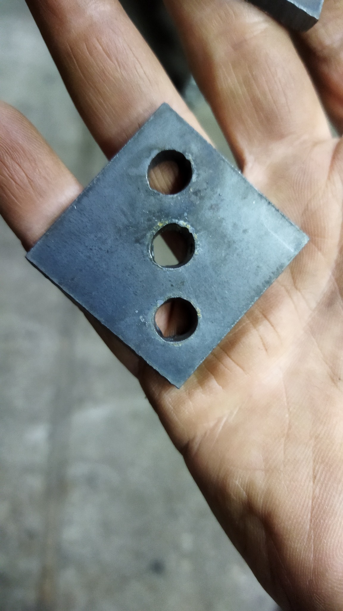 Metalowy element w kształcie rombu z trzema okrągłymi otworami, trzymany w dłoni.