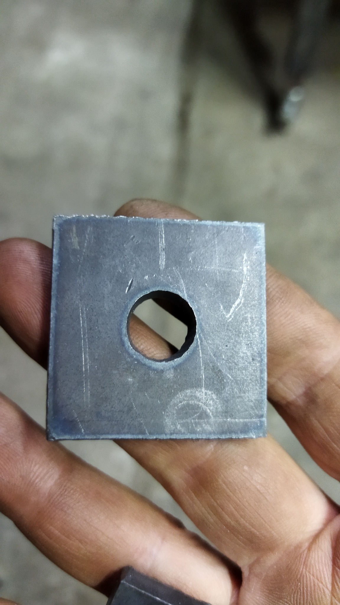 Metalowy kwadrat z okrągłym otworem pośrodku, trzymany w dłoniach, widoczne ślady obróbki i zarysowania.