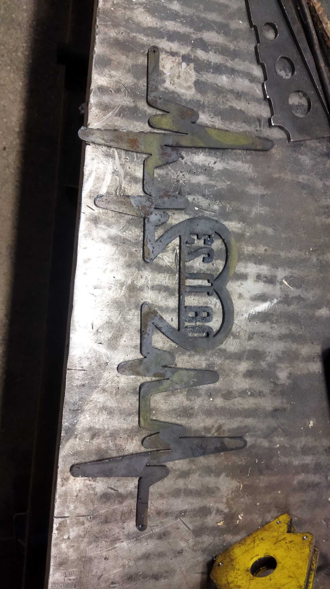 Metalowy element wycięty w kształcie zapisu EKG z napisem 'ESTIBAI' w środku, leżący na brudnym, metalowym stole warsztatowym.