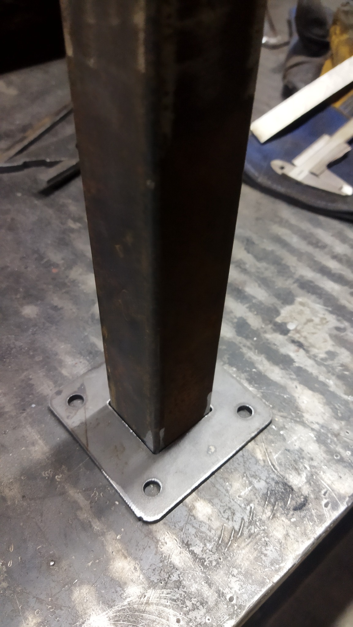 Metalowy profil kwadratowy przyspawany do blachy z otworami, widoczne ślady obróbki na powierzchni blatu roboczego.