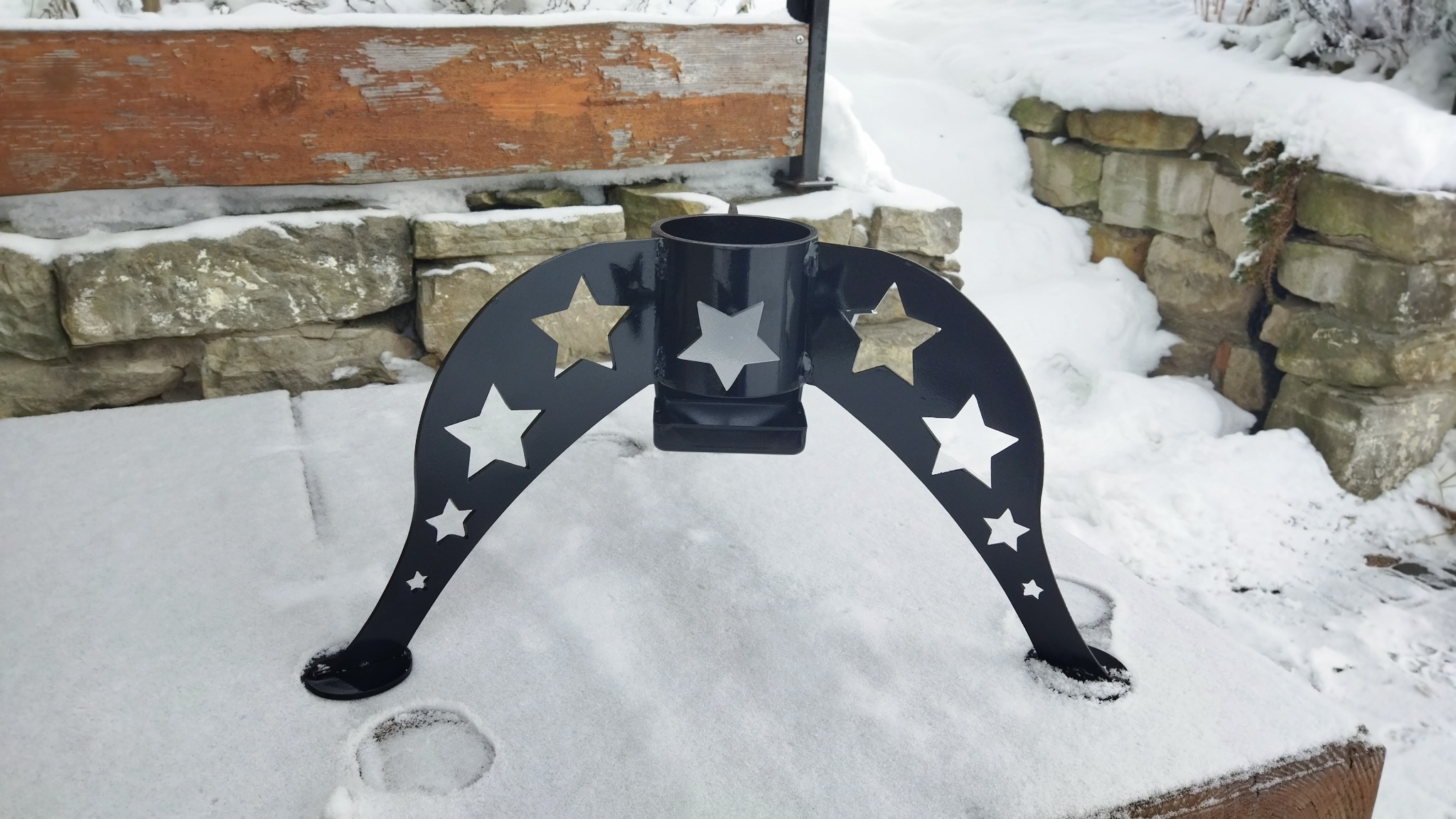 Czarny, metalowy stojak na choinkę z wyciętymi gwiazdami, umieszczony na śniegu przy kamiennym murku i drewnianej ławce.